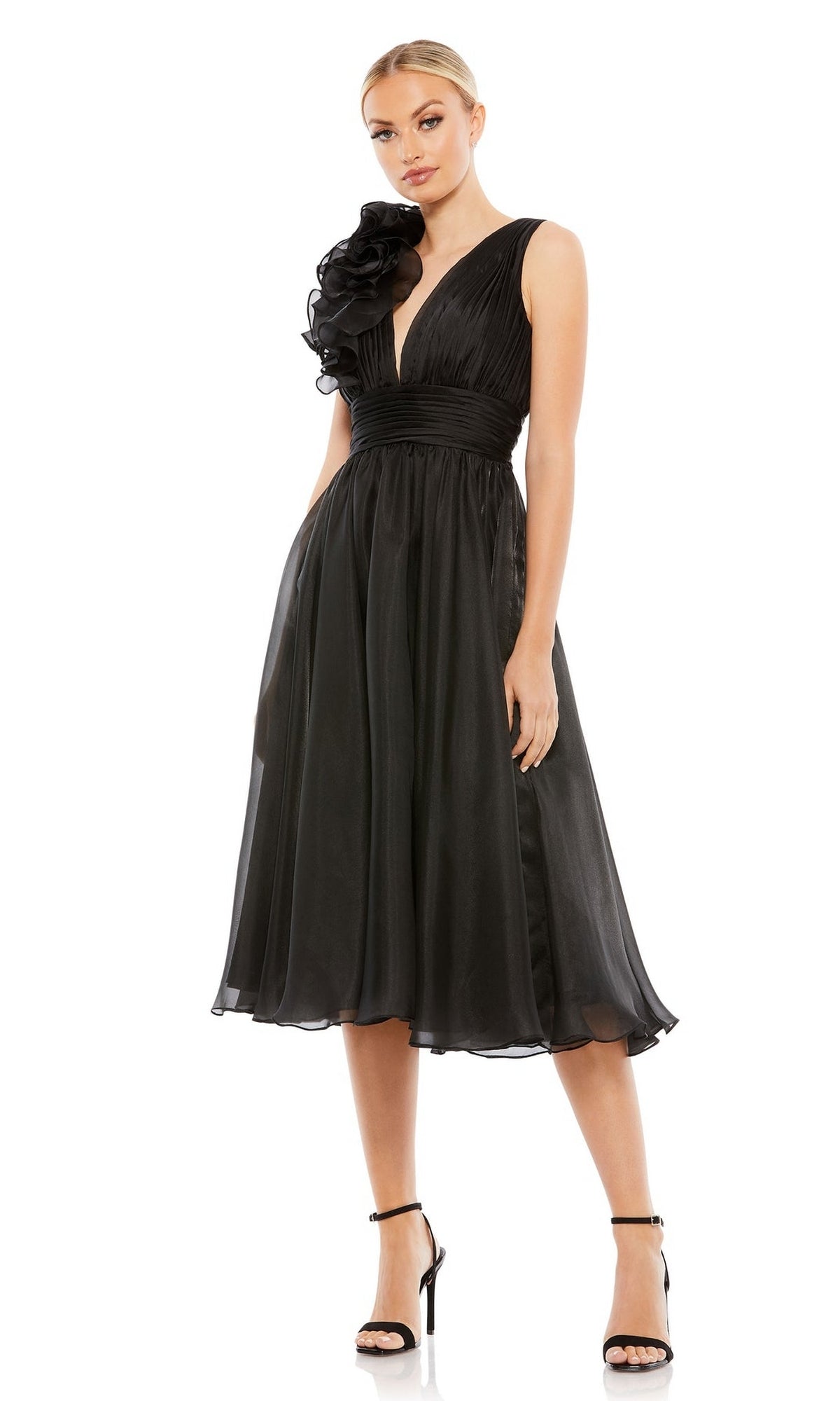 A woman wears the Mac Duggal Short Wedding Guest Dress 70165—a sleeveless black knee-length dress featuring a deep V-neck and large ruffle on one shoulder—paired with black high-heeled sandals.